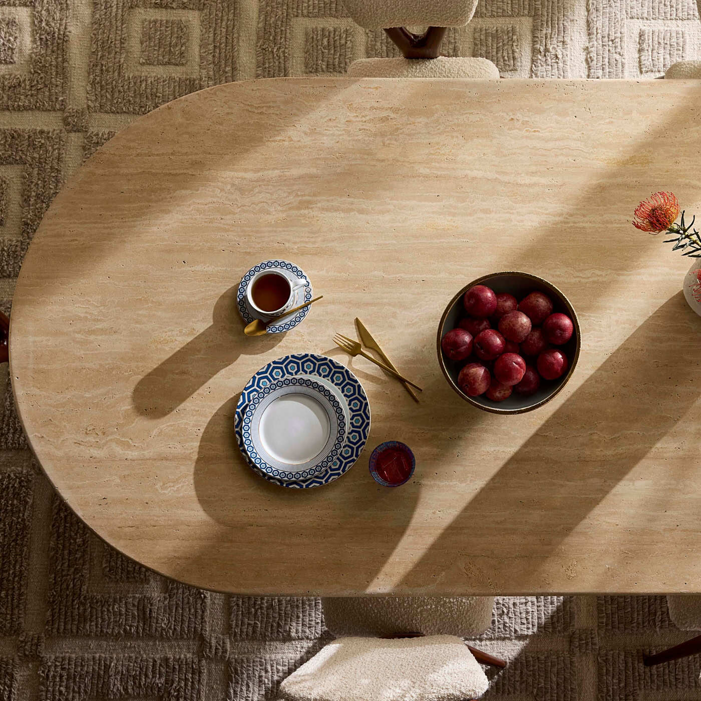 Wooden table with a cup, saucer, bowl of apples, and other items on a textured floor.