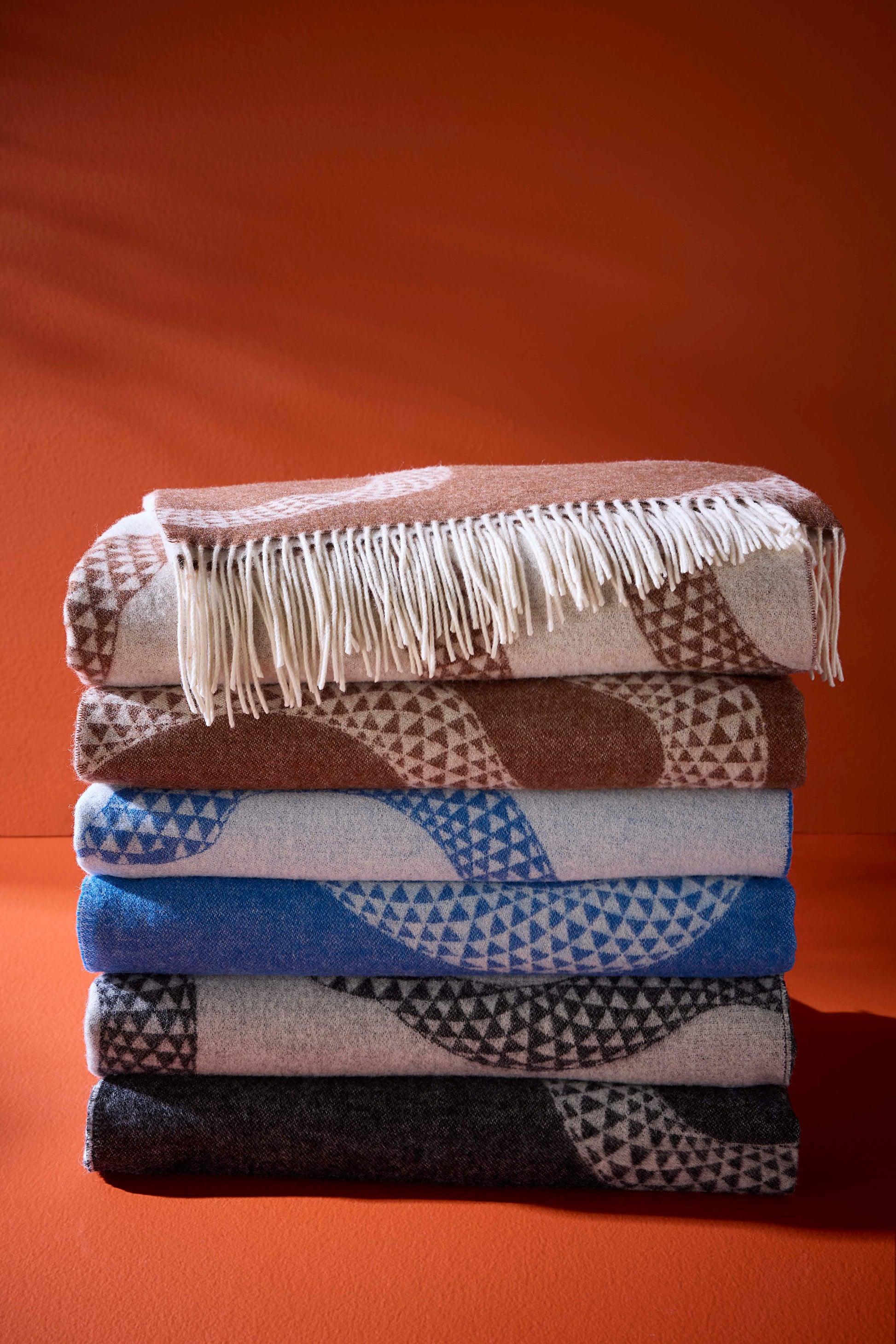 A stack of five folded woven blankets with geometric patterns and fringe, displayed against a solid terracotta orange background.