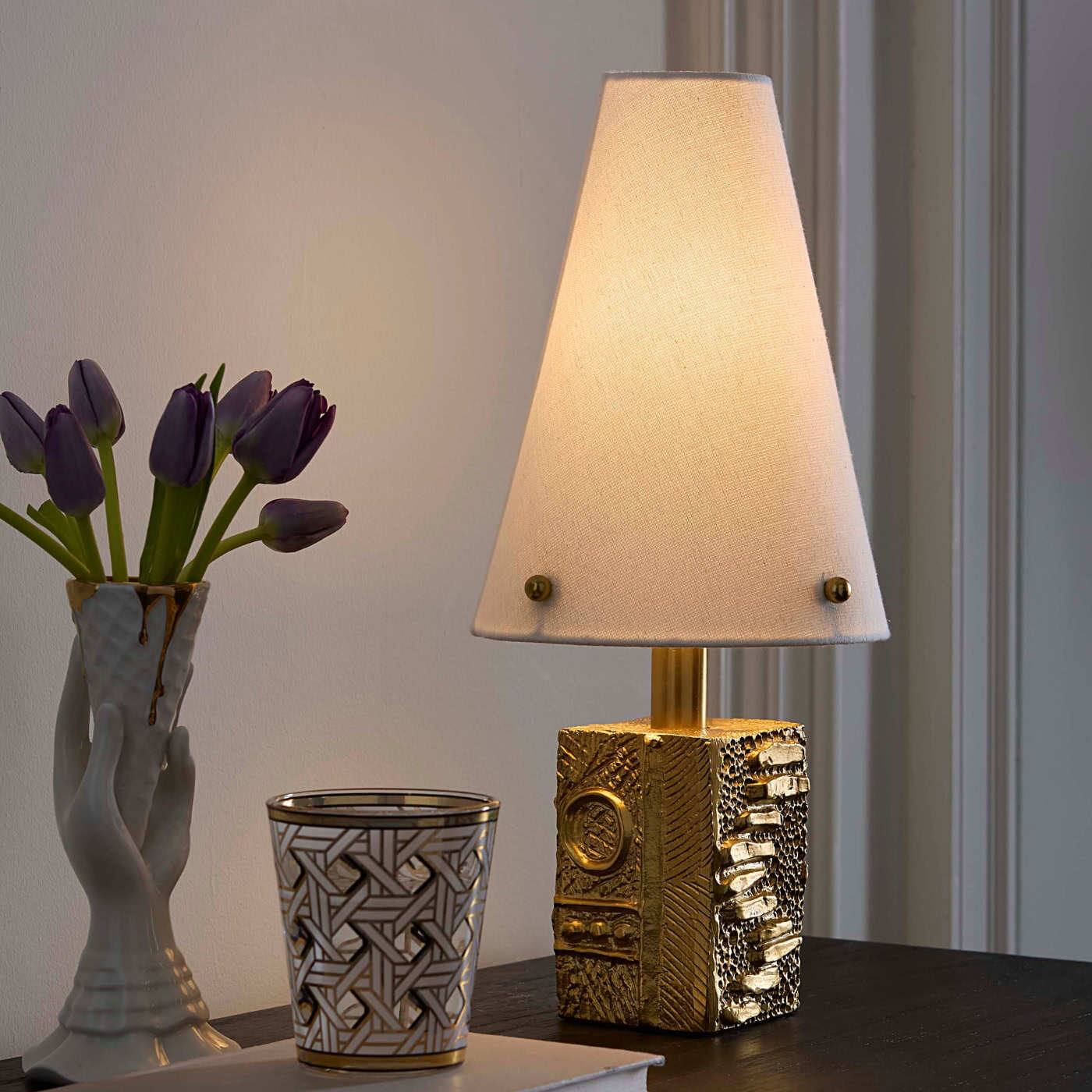 Decorative lamp with textured base on a table next to a vase with purple tulips and a patterned glass.