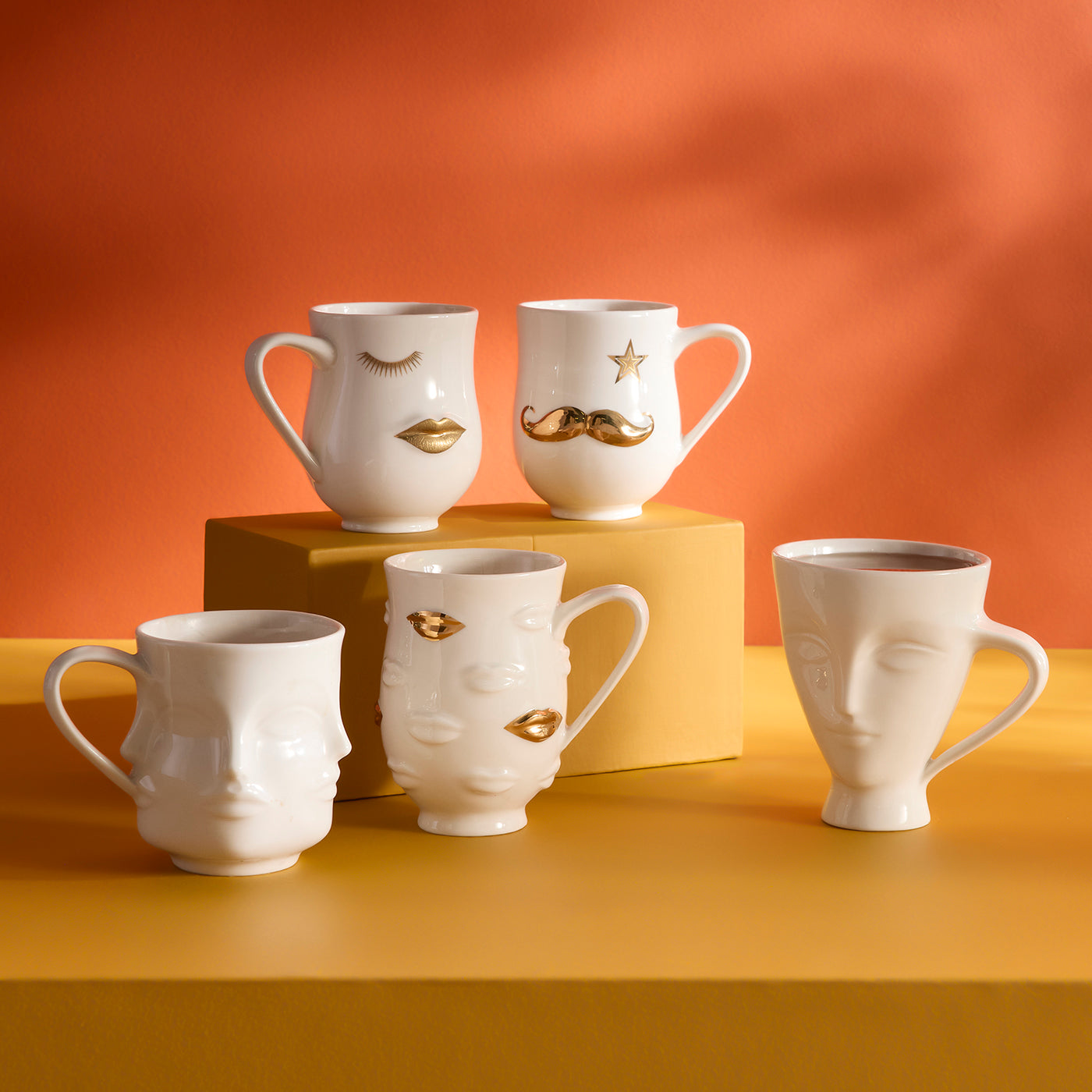 Five white ceramic mugs with sculptural face designs and gold decorative accents, arranged on a yellow surface with an orange backdrop.