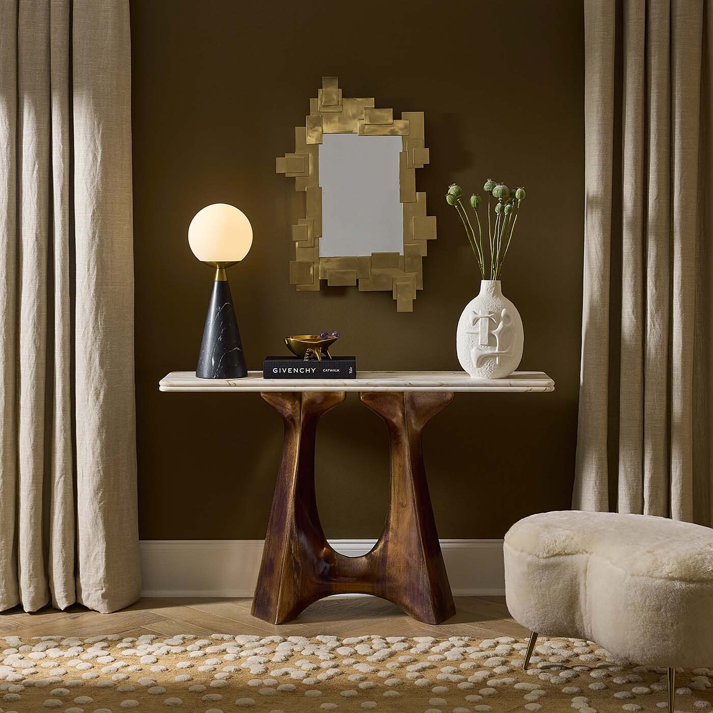 A styled interior scene showing a console table with a sculptural dark wood base and a light stone top, set against a dark brown wall with curtains, a gold-framed mirror, a lit table lamp, a white vase with green stems, and a cream upholstered ottoman.