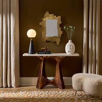 A styled interior scene showing a console table with a sculptural dark wood base and a light stone top, set against a dark brown wall with curtains, a gold-framed mirror, a lit table lamp, a white vase with green stems, and a cream upholstered ottoman.