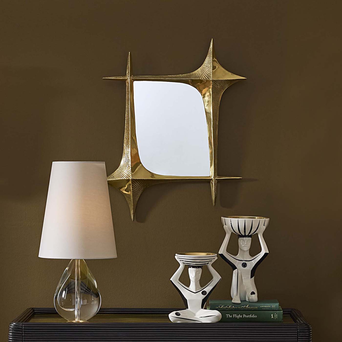 Decorative mirror with geometric design above a shelf with a lamp, books, and sculptures against a brown wall.
