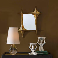 Decorative mirror with geometric design above a shelf with a lamp, books, and sculptures against a brown wall.