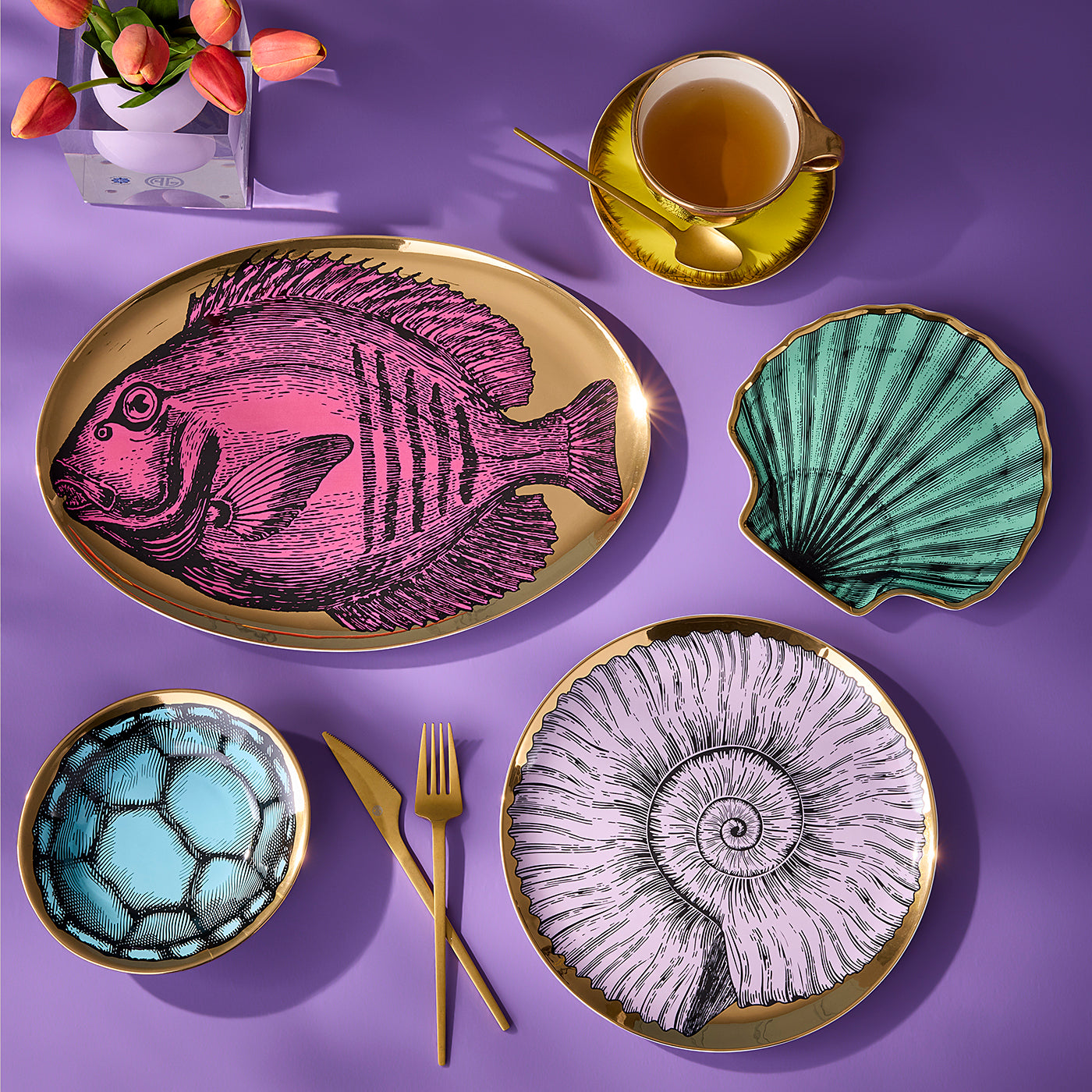 A styled flat-lay of a dinnerware set on a purple background, featuring a large oval platter with a pink fish illustration, a scallop-shaped dish in teal, a bowl with a blue soccer pattern, and two round plates with gold-leaf-edged nautilus and spiral designs. Gold cutlery, a yellow teacup, and a vase of tulips are arranged around them.