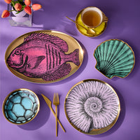 A styled flat-lay of a dinnerware set on a purple background, featuring a large oval platter with a pink fish illustration, a scallop-shaped dish in teal, a bowl with a blue soccer pattern, and two round plates with gold-leaf-edged nautilus and spiral designs. Gold cutlery, a yellow teacup, and a vase of tulips are arranged around them.