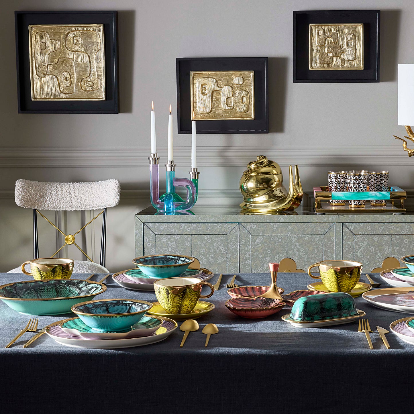 A styled dining table set with gold and turquoise ceramic tableware, including teacups, plates, and butter dishes, alongside matching gold and turquoise candlesticks and a gold teapot on a mirrored sideboard. Three framed abstract gold artworks hang on a gray wall above.