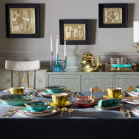 A styled dining table set with gold and turquoise ceramic tableware, including teacups, plates, and butter dishes, alongside matching gold and turquoise candlesticks and a gold teapot on a mirrored sideboard. Three framed abstract gold artworks hang on a gray wall above.