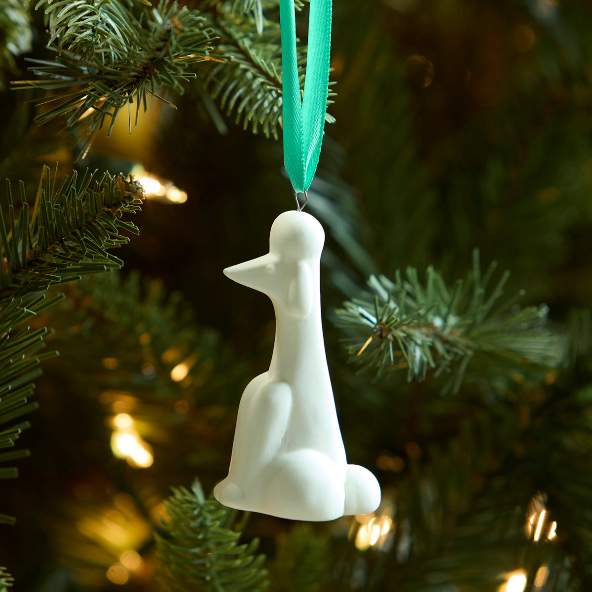 A white ceramic penguin Christmas ornament hanging from a mint green ribbon on a decorated Christmas tree with warm lights.