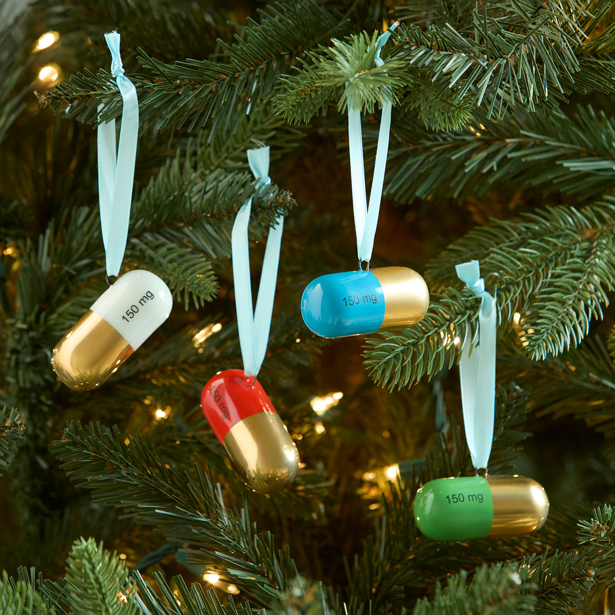 Four capsule-shaped Christmas ornaments hanging from a green Christmas tree with warm string lights. The capsules are white, blue, red, and green, each with a white ribbon loop and printed text reading '150 mg'.