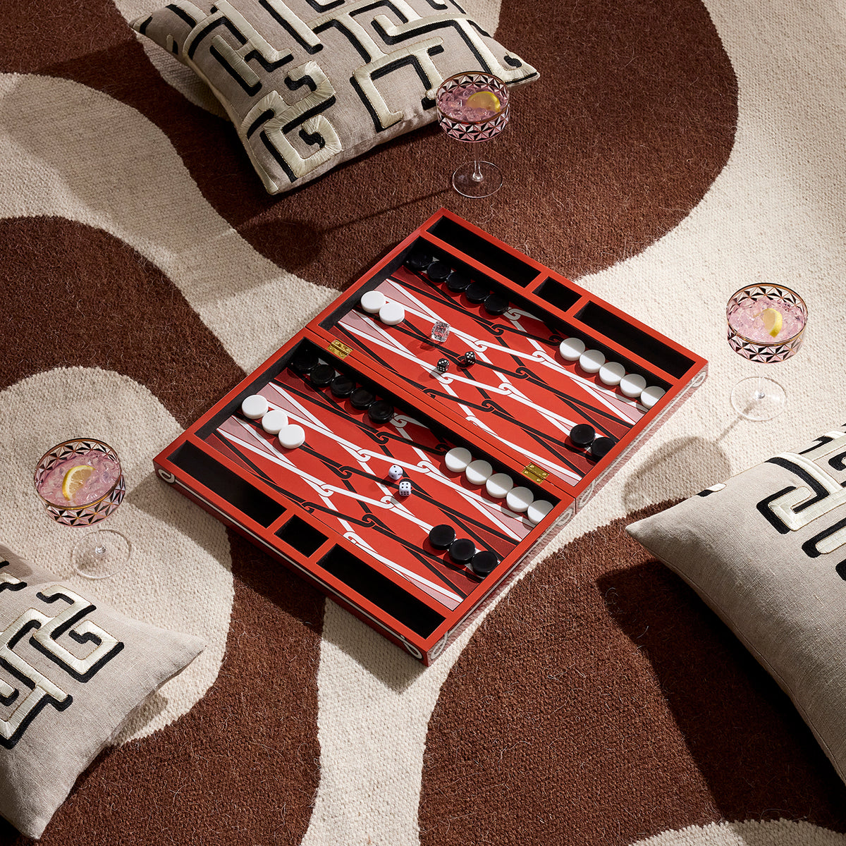 A red wooden backgammon board with white and black checkers is arranged on a patterned brown and beige rug, surrounded by decorative pillows and two wine glasses.