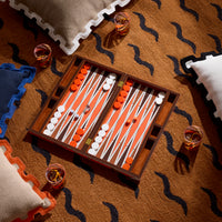 An open wooden backgammon set with orange and white playing pieces arranged on a patterned brown rug, surrounded by pillows and two glasses of amber liquid.