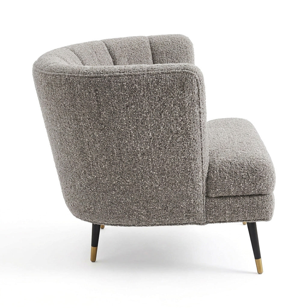 A modern curved armchair upholstered in a textured gray bouclé fabric, featuring tapered black legs with gold metal tips, shown against a plain white background.