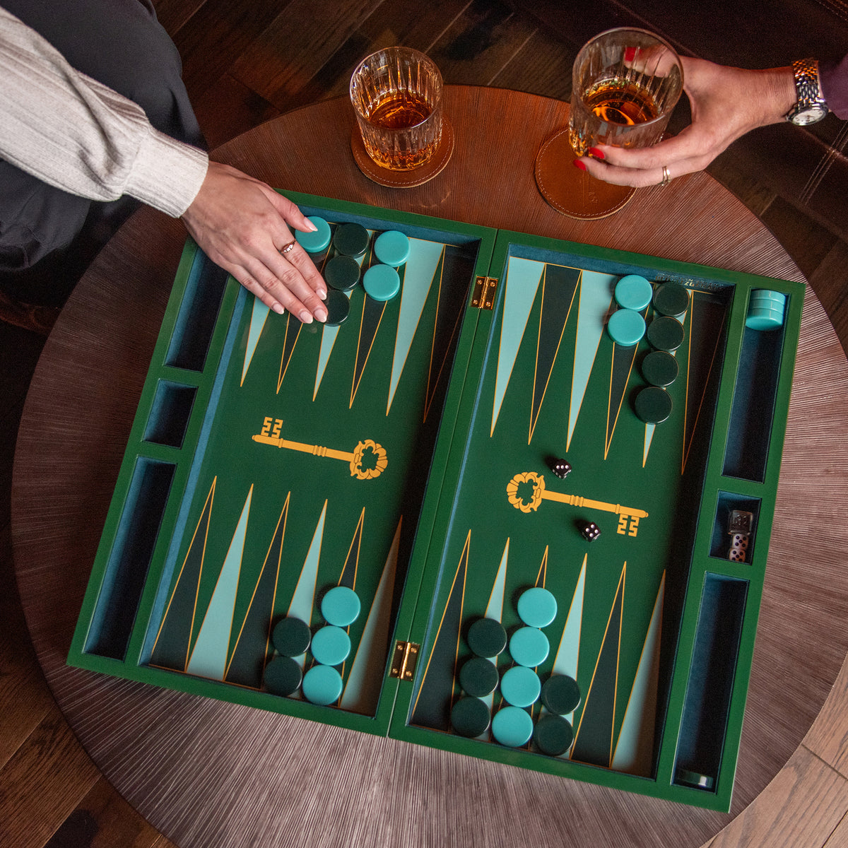 A wooden backgammon board set on a round wooden table, with two people's hands holding drinks above it. The board is green with gold lines and teal and dark green checkers.