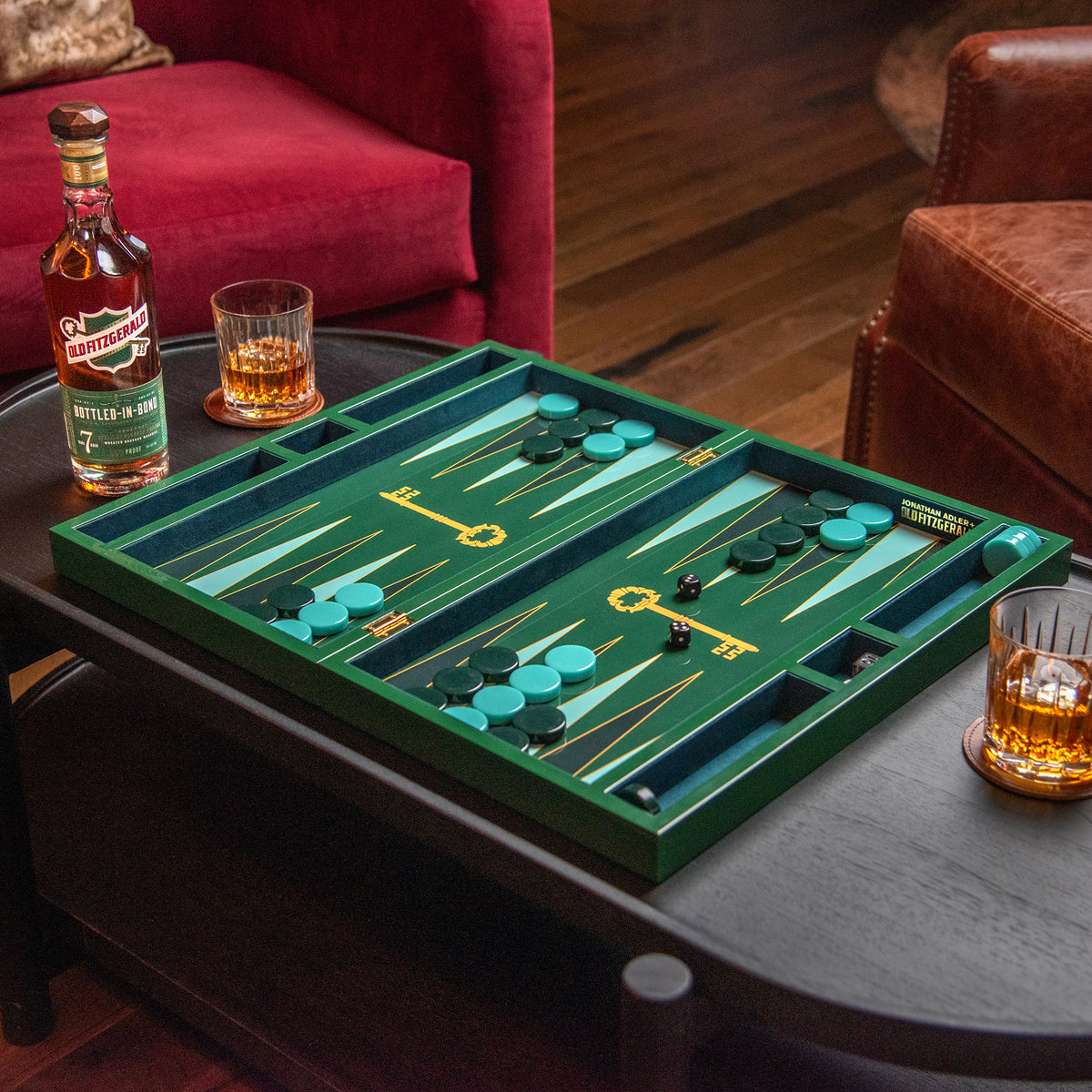 A green backgammon board set with teal and gold playing pieces arranged on a dark wooden coffee table in a living room setting, with a bottle of whiskey and two glasses of amber liquor nearby.