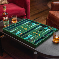 A green backgammon board set with teal and gold playing pieces arranged on a dark wooden coffee table in a living room setting, with a bottle of whiskey and two glasses of amber liquor nearby.