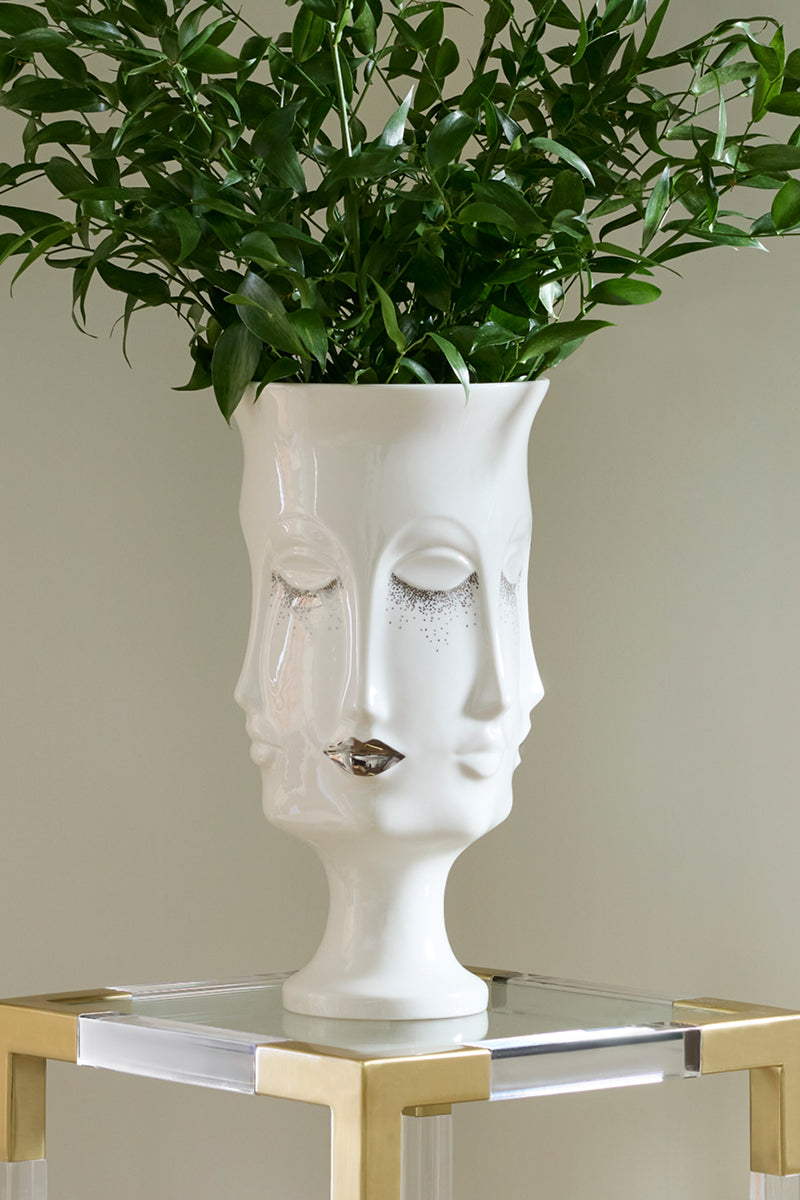 A white ceramic planter shaped like a stylized female face with closed eyes and metallic silver glitter details, filled with green foliage, displayed on a gold-framed glass side table against a neutral beige wall.