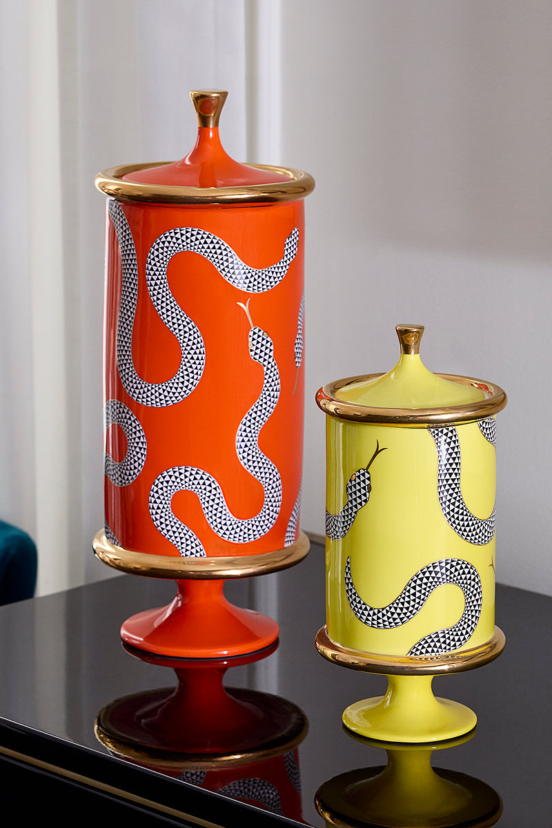 Two decorative ceramic lidded jars with pedestal bases, one orange and one yellow, both featuring a black-and-white spotted snake pattern and gold trim, displayed on a reflective black surface.
