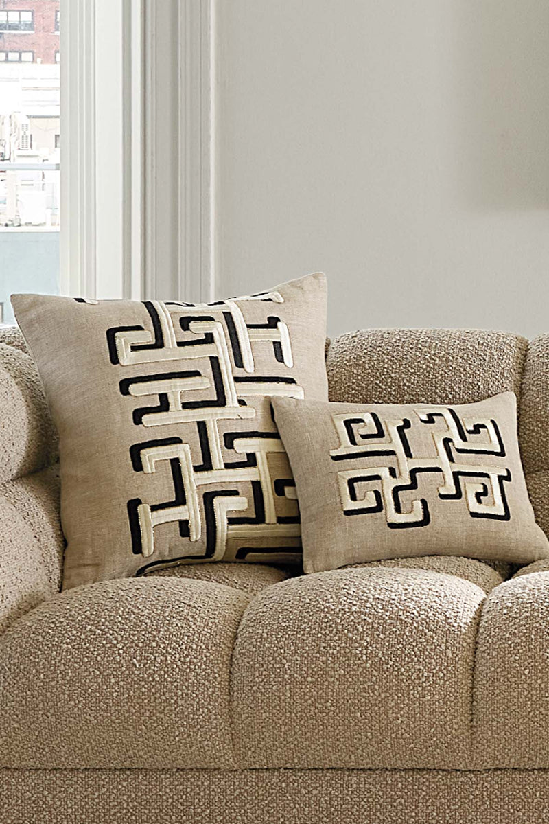 Two beige throw pillows with black geometric Greek key patterns resting on a textured beige boucle sofa in front of a window.