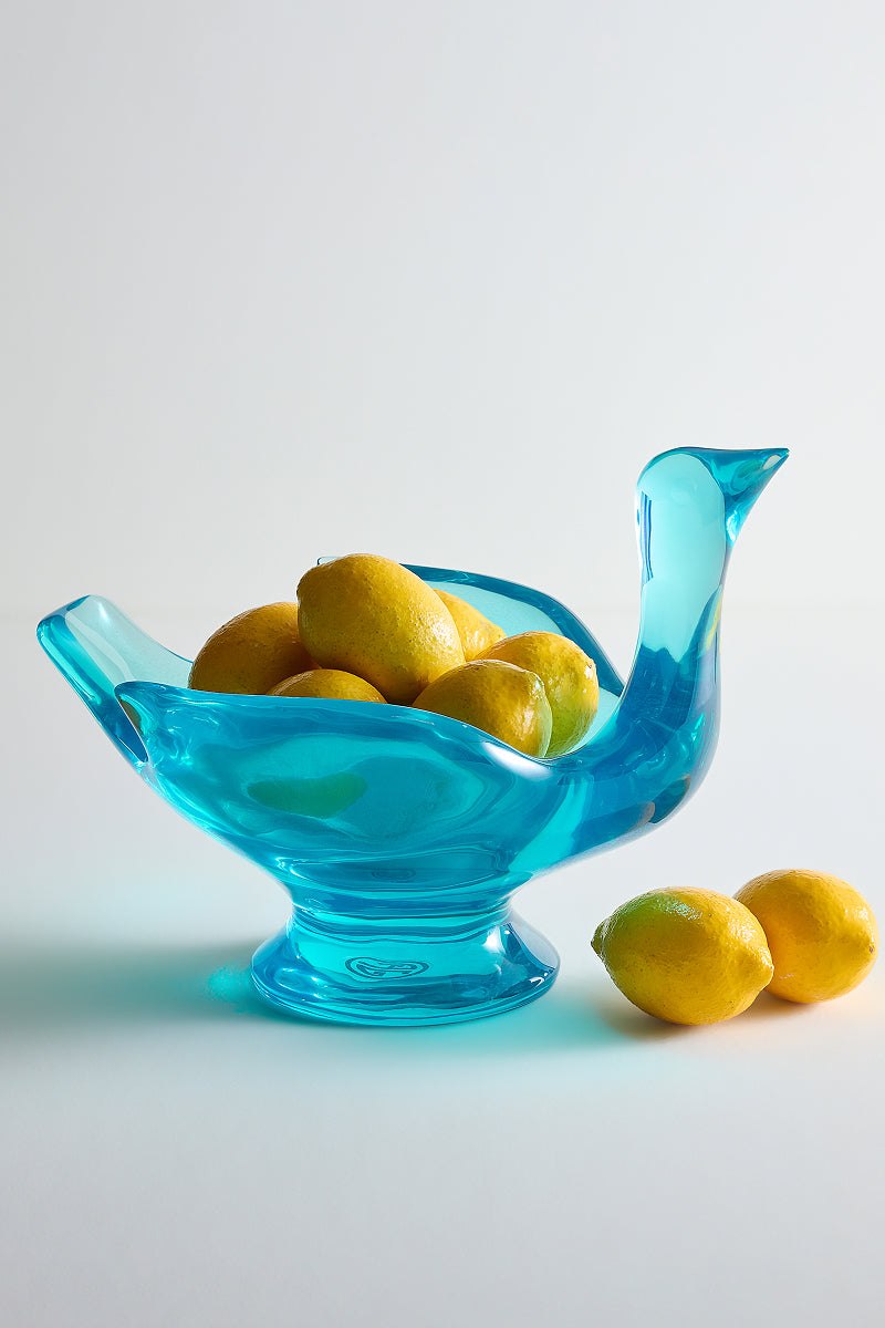 A translucent turquoise blue bird-shaped glass bowl filled with yellow lemons, with two additional lemons resting beside it on a clean white studio background.