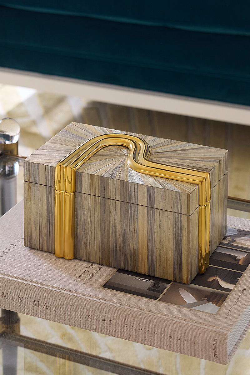 A rectangular decorative box with a striped wood-grain pattern in beige, brown, and gray, accented by a prominent curved gold metal handle and gold trim, resting on a book.