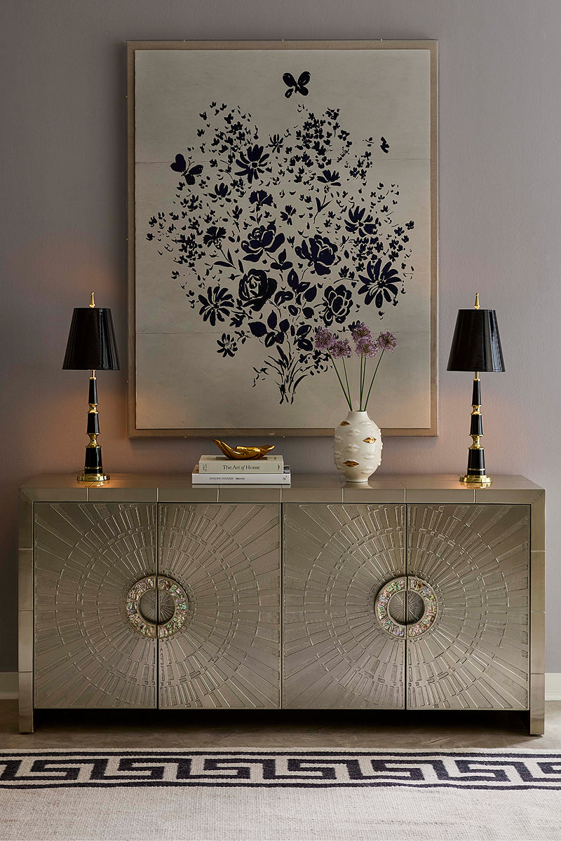 A styled interior scene featuring a large framed black floral artwork on a light gray wall above a metallic silver credenza, with two black table lamps, a small vase of flowers, and a patterned rug.