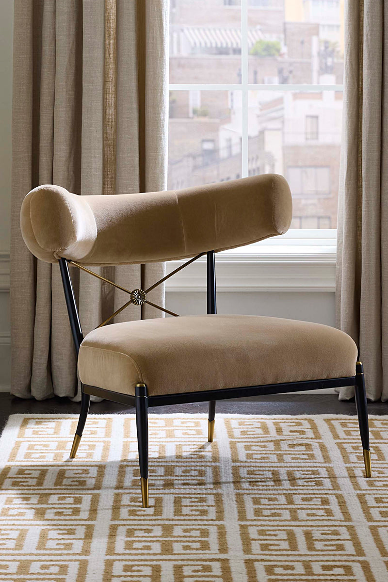 A modern lounge chair with a curved beige velvet backrest and seat, supported by black metal legs with gold-tone tips, set on a patterned rug in front of a window with curtains.