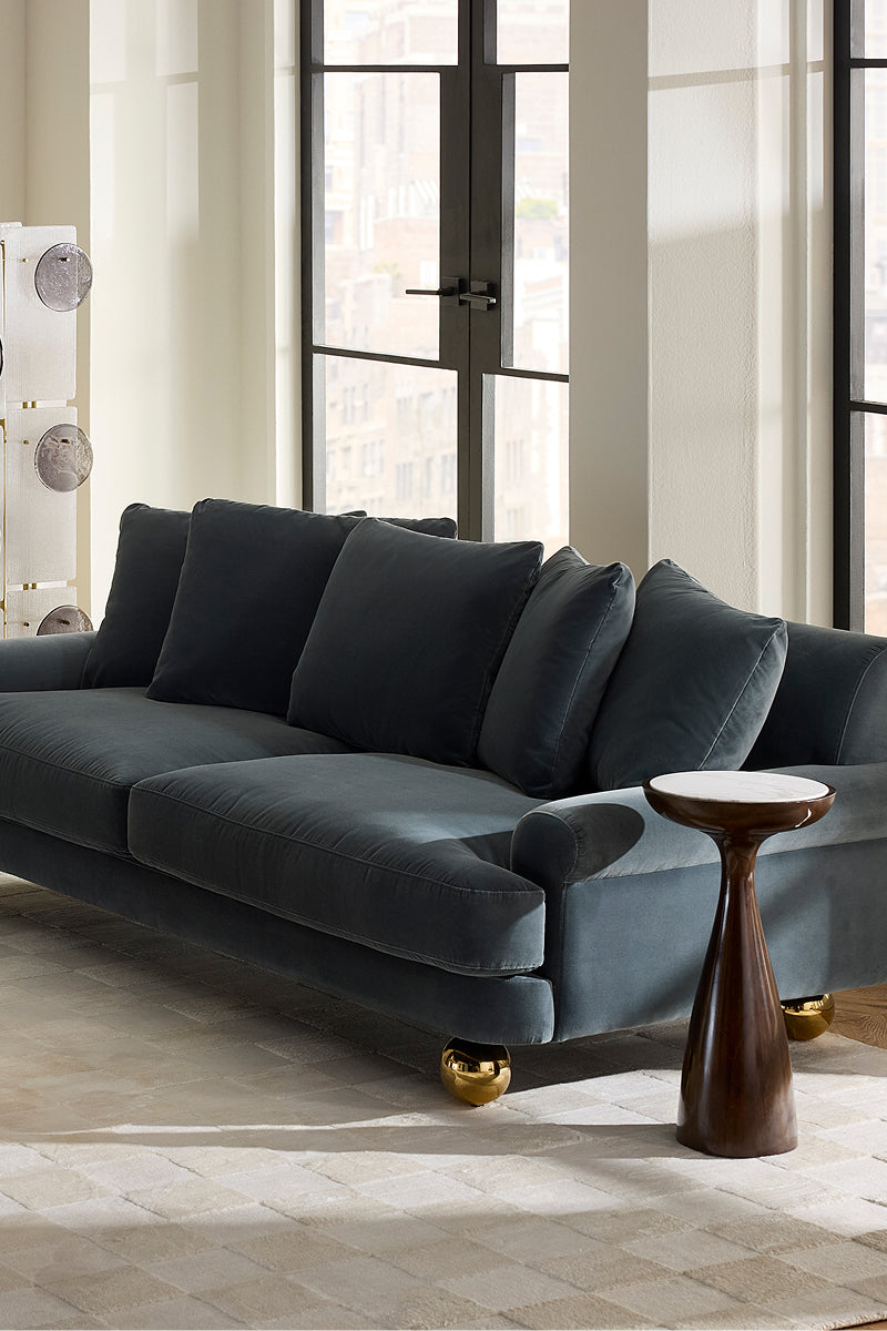 A modern dark blue velvet sofa with gold ball feet, styled in a bright room with a small wooden side table next to it.