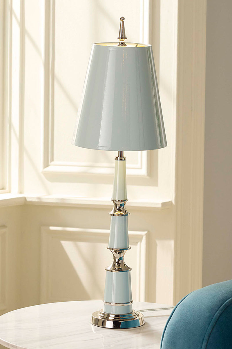 A table lamp with a light blue-gray shade and matching base, accented with polished gold-toned metal and a white ceramic neck, displayed on a white marble table in a sunlit room.