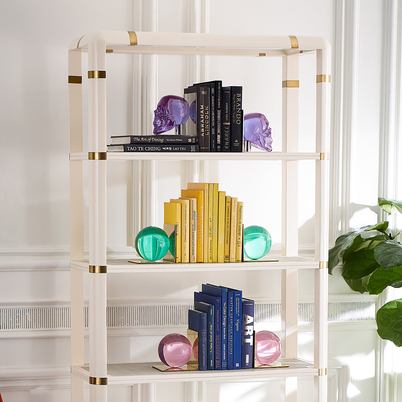 A tall white bookshelf with gold accents, styled with books, decorative crystal-like spheres, and bookends in a bright room with a window and plant.