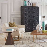 A styled interior scene featuring a tall black cabinet with embossed square medallion panels, positioned next to a cream boucle armchair and a wooden coffee table in a bright room with white paneled walls.