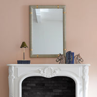 A rectangular wall mirror with a textured beige linen frame and gold corner accents, mounted on a dusty pink wall above a white fireplace mantel styled with books and horse figurines.