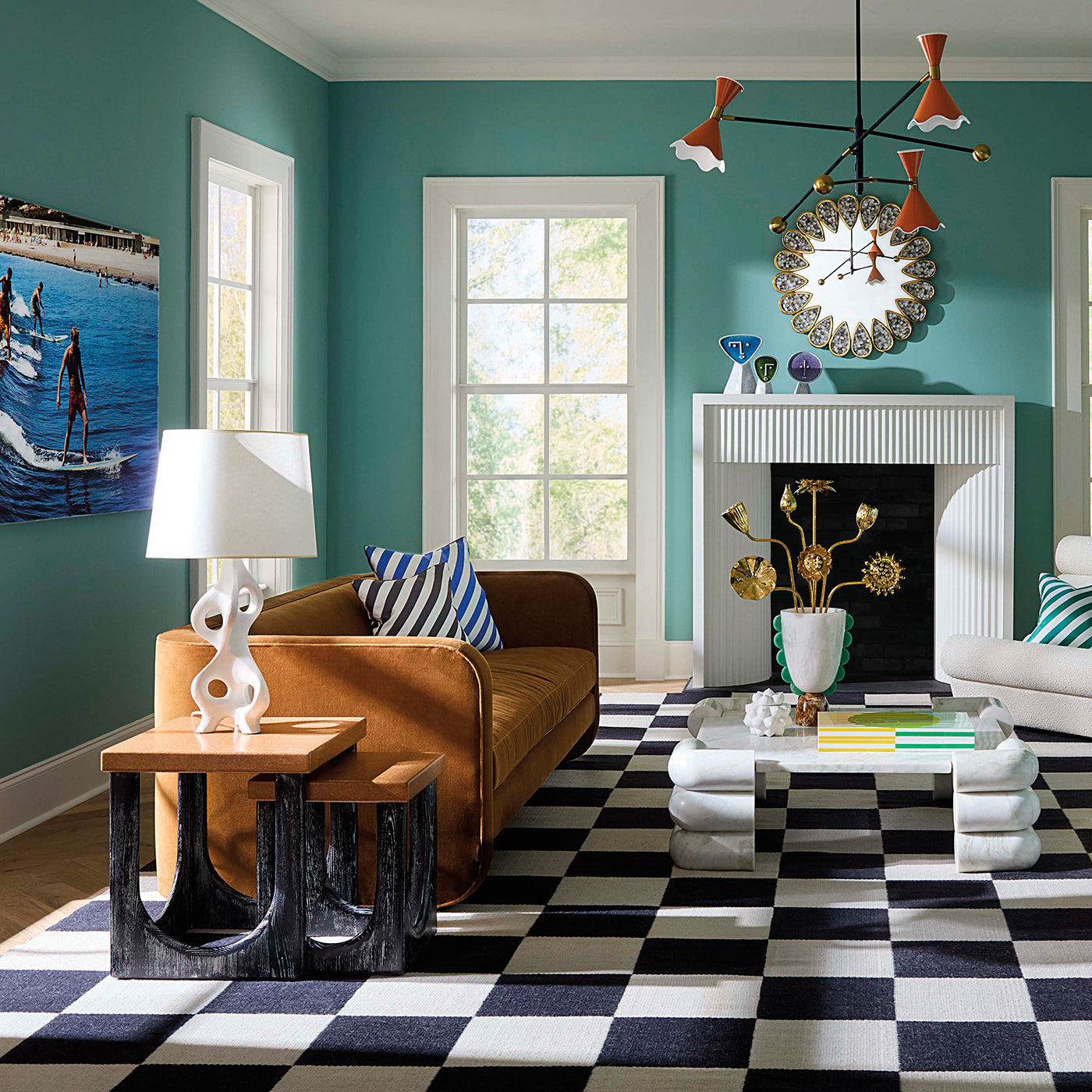 A styled living room with teal walls, a brown leather sofa, a black-and-white checkered rug, a white fireplace, and multiple decorative objects including a lamp, side table, wall art, and a chandelier.