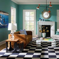A styled living room with teal walls, a brown leather sofa, a black-and-white checkered rug, a white fireplace, and multiple decorative objects including a lamp, side table, wall art, and a chandelier.