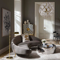 A modern curved gray velvet sofa in a styled living room with a gold floor lamp, a round mirrored coffee table, a white floral chandelier, and dark gray walls.