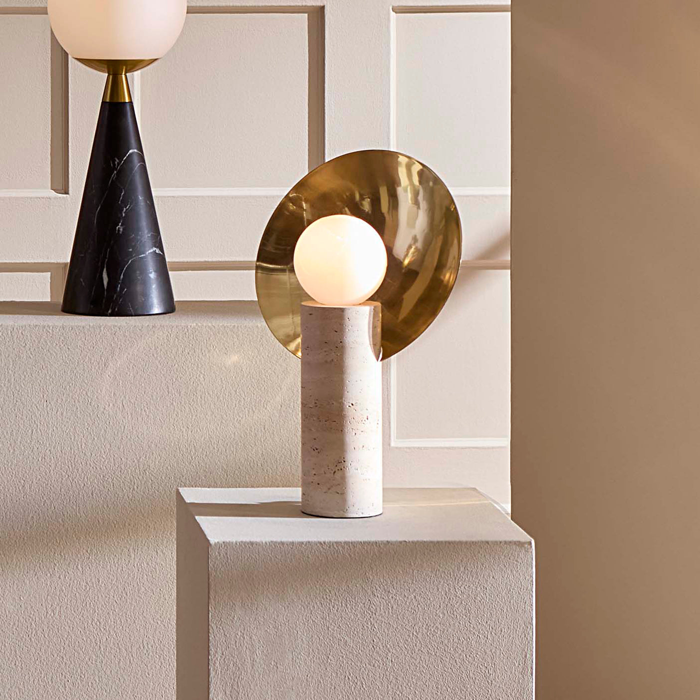 A modern table lamp with a spherical white globe, a textured beige cylindrical base, and a large polished gold circular backplate, displayed on a white pedestal against a beige paneled wall.