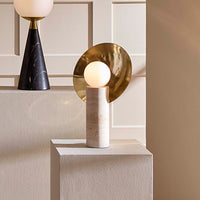 A modern table lamp with a spherical white globe, a textured beige cylindrical base, and a large polished gold circular backplate, displayed on a white pedestal against a beige paneled wall.