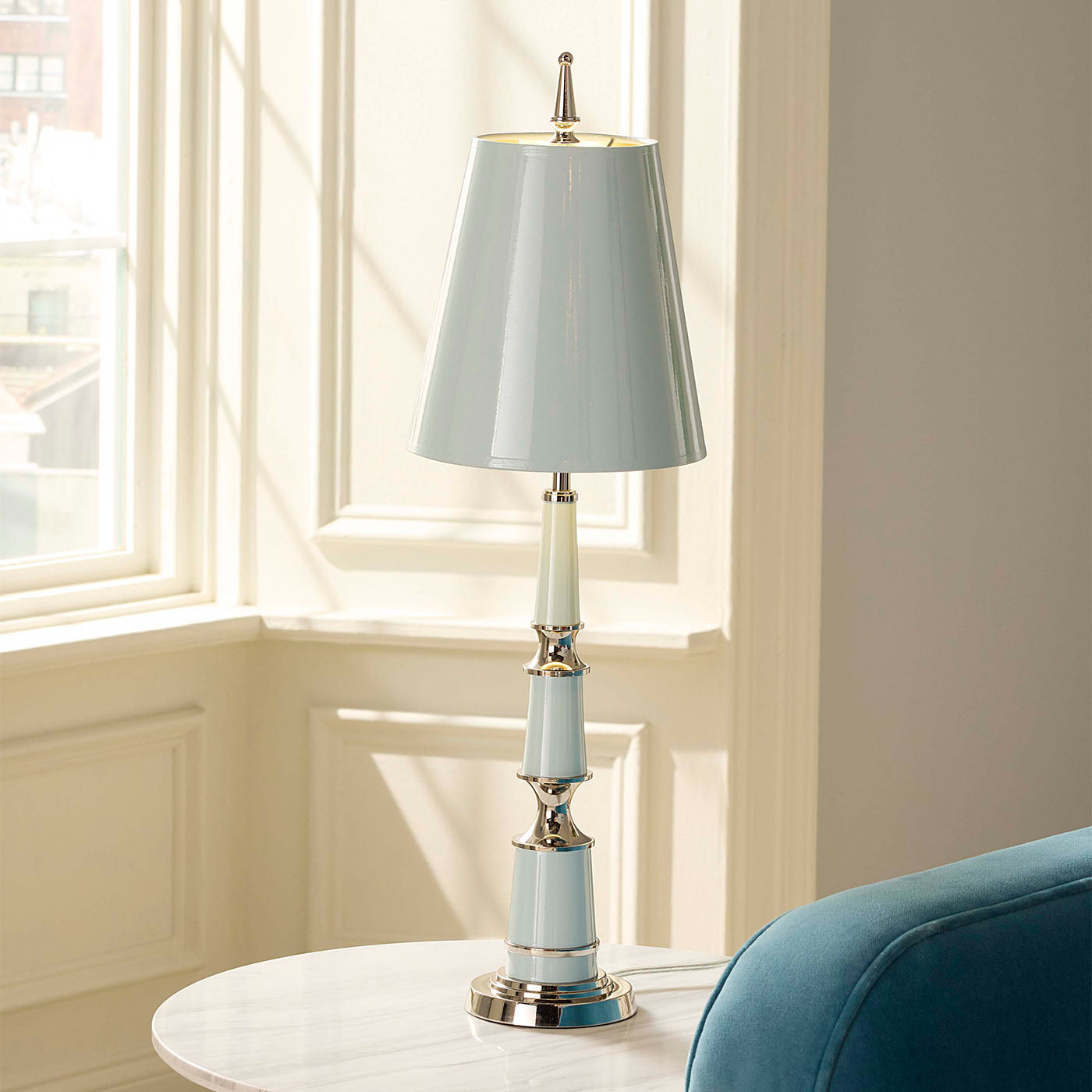 Light blue table lamp with silver base on a white surface near a window.