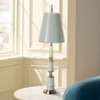 Light blue table lamp with silver base on a white surface near a window.