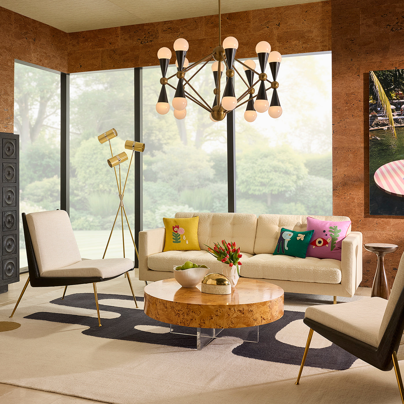 A modern living room scene featuring a cream-colored sofa with colorful decorative pillows, a round burl wood coffee table, a black and cream accent chair, and a multi-light chandelier hanging from a central ceiling.