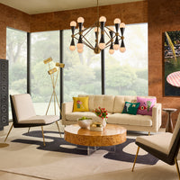 A modern living room scene featuring a cream-colored sofa with colorful decorative pillows, a round burl wood coffee table, a black and cream accent chair, and a multi-light chandelier hanging from a central ceiling.