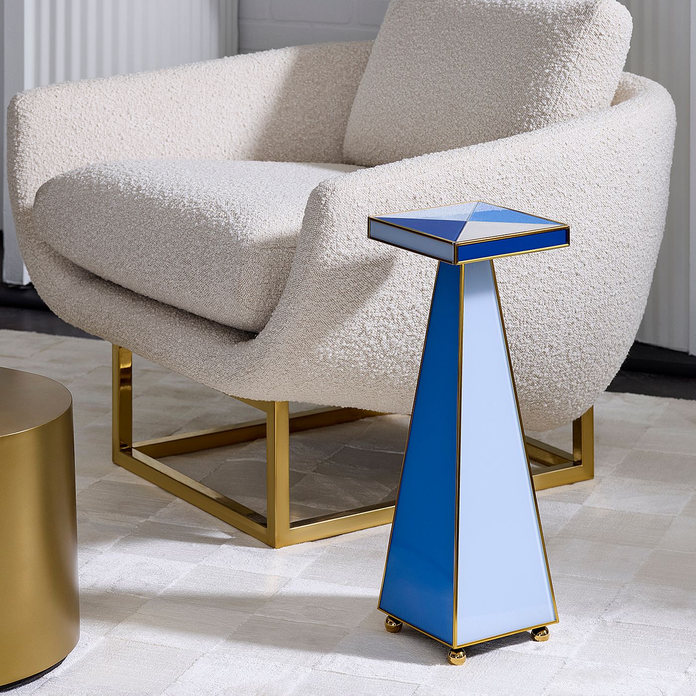 A modern geometric side table with a blue and white triangular design and gold brass trim, styled next to a cream bouclé armchair with a gold base.