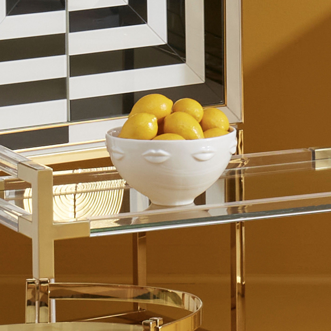 Bowl of lemons on a glass table with gold legs against a striped wall.