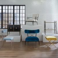 Modern furniture pieces including a blue chair, yellow stool, and black cabinet in a room with large windows and white brick walls.