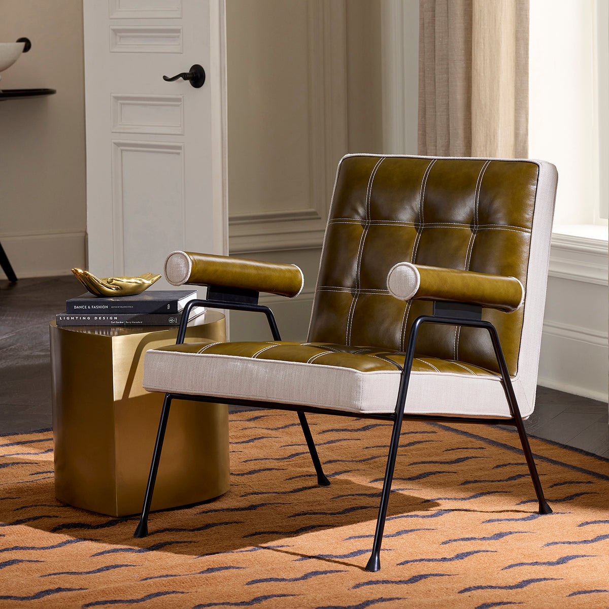 A modern accent chair with olive green tufted leather and cream fabric upholstery, supported by black metal legs, styled in a room next to a gold side table.