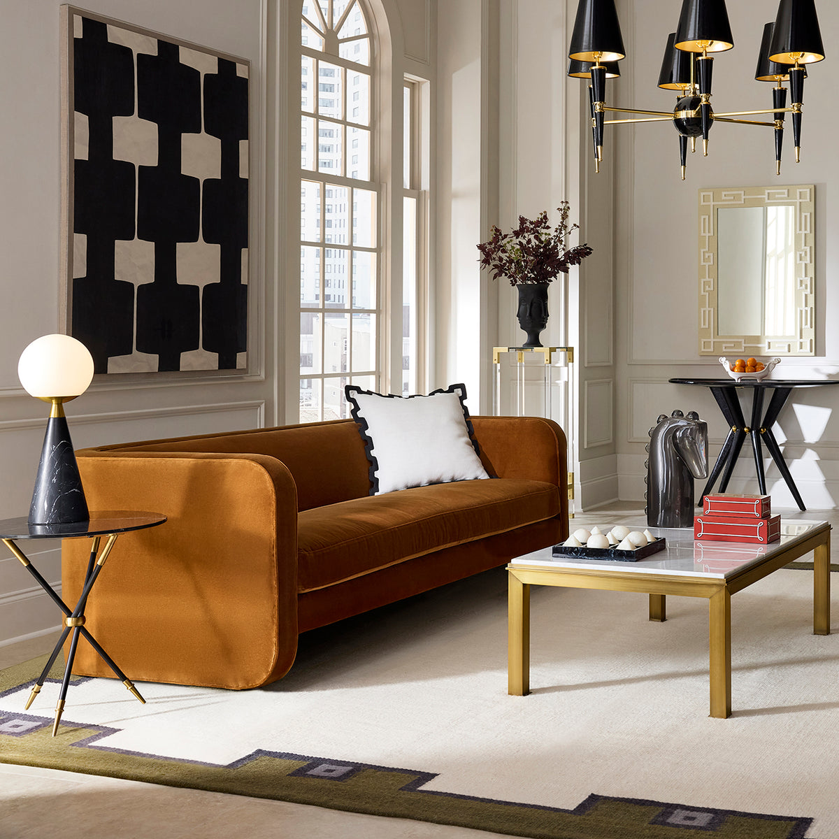 A modern living room featuring a camel-brown velvet sofa with a white accent pillow, a marble-top coffee table with gold legs, a black side table with a globe lamp, and a large black and white geometric wall art piece.