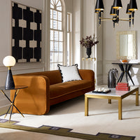 A modern living room featuring a camel-brown velvet sofa with a white accent pillow, a marble-top coffee table with gold legs, a black side table with a globe lamp, and a large black and white geometric wall art piece.
