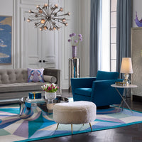 A styled living room interior featuring a blue tufted armchair, a gray sofa, a white fluffy ottoman, a geometric blue and purple area rug, a chandelier, and a side table with a lamp.