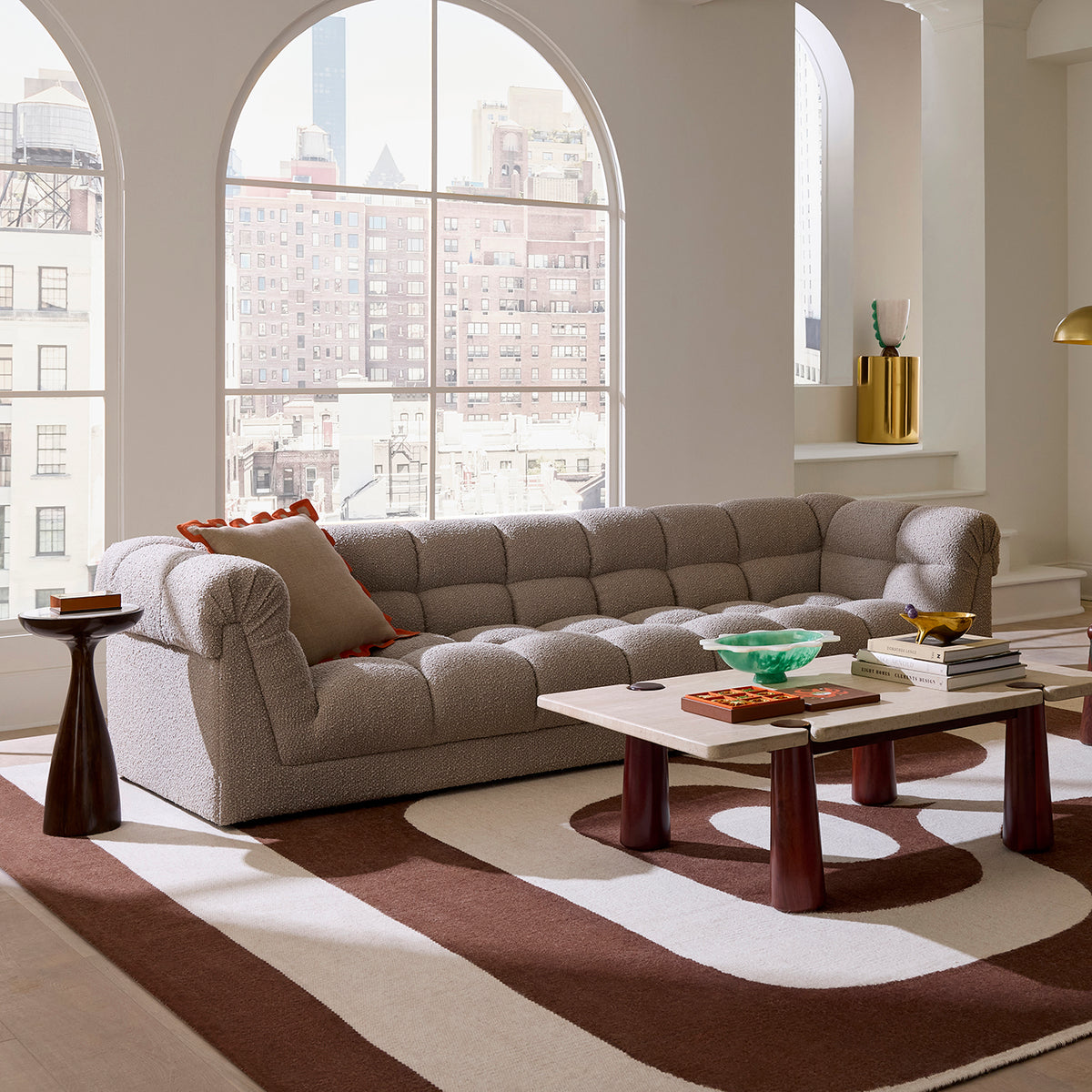 A modern tufted sofa in light beige upholstery, styled in a bright, contemporary living room with arched windows, a striped area rug, and a wooden coffee table.
