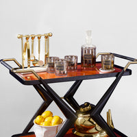 A luxury bar cart with a black and reddish-brown wood frame, styled with gold-toned glassware, a decanter, and lemons on a light gray background.